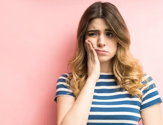 Woman in striped shirt touching face against pink background, suggesting dental concern or discomfort.