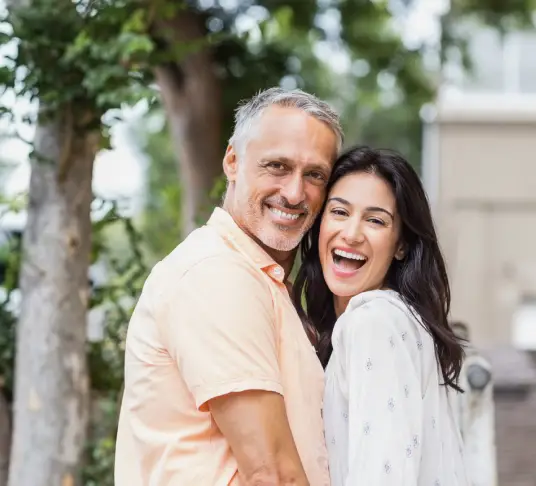 Smiling mature couple embracing outdoors near trees and building, periodontal health context.